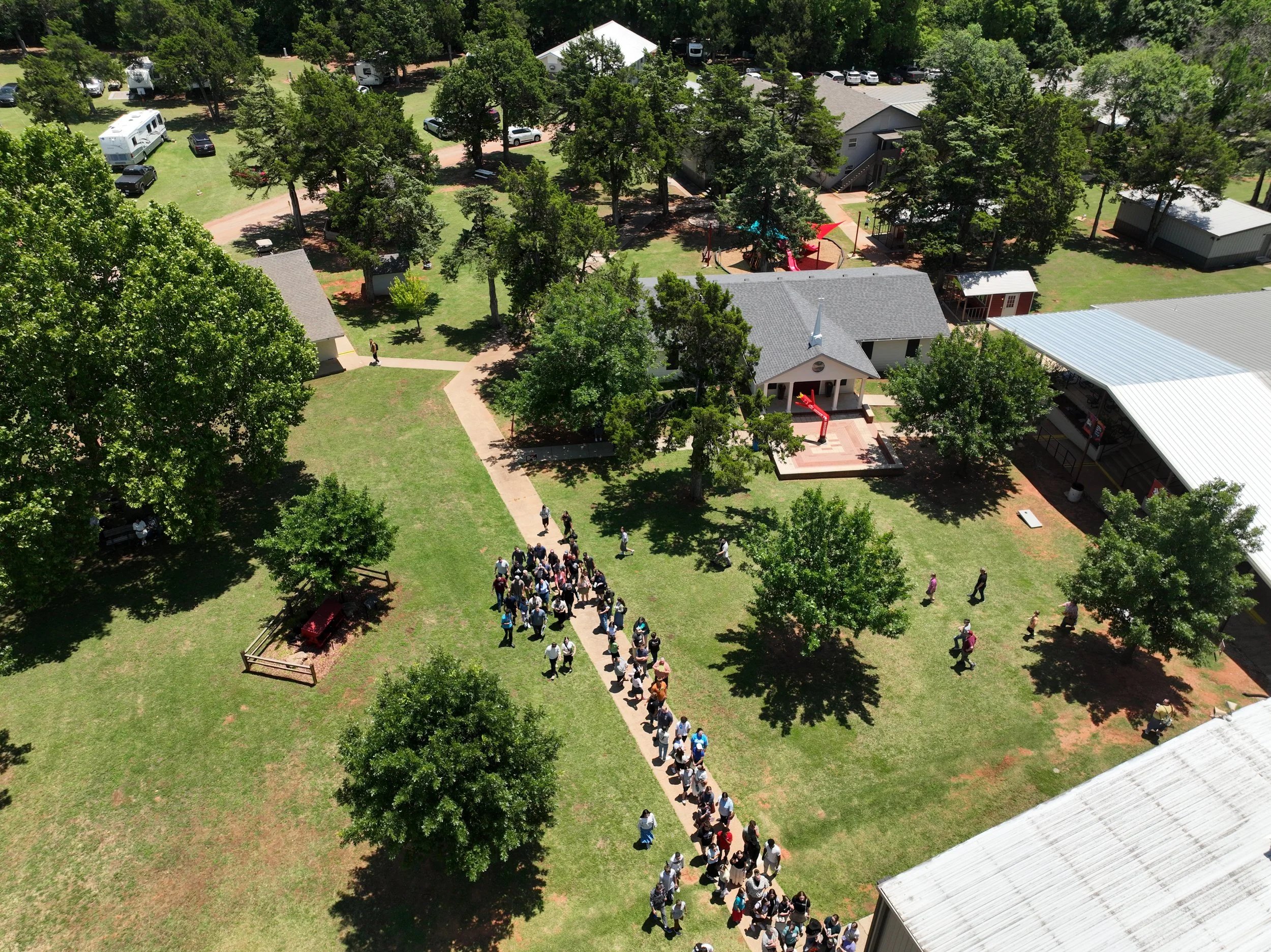 Aerial view of a park with a line of people walking on a path, surrounded by trees, small buildings, and recreational areas.