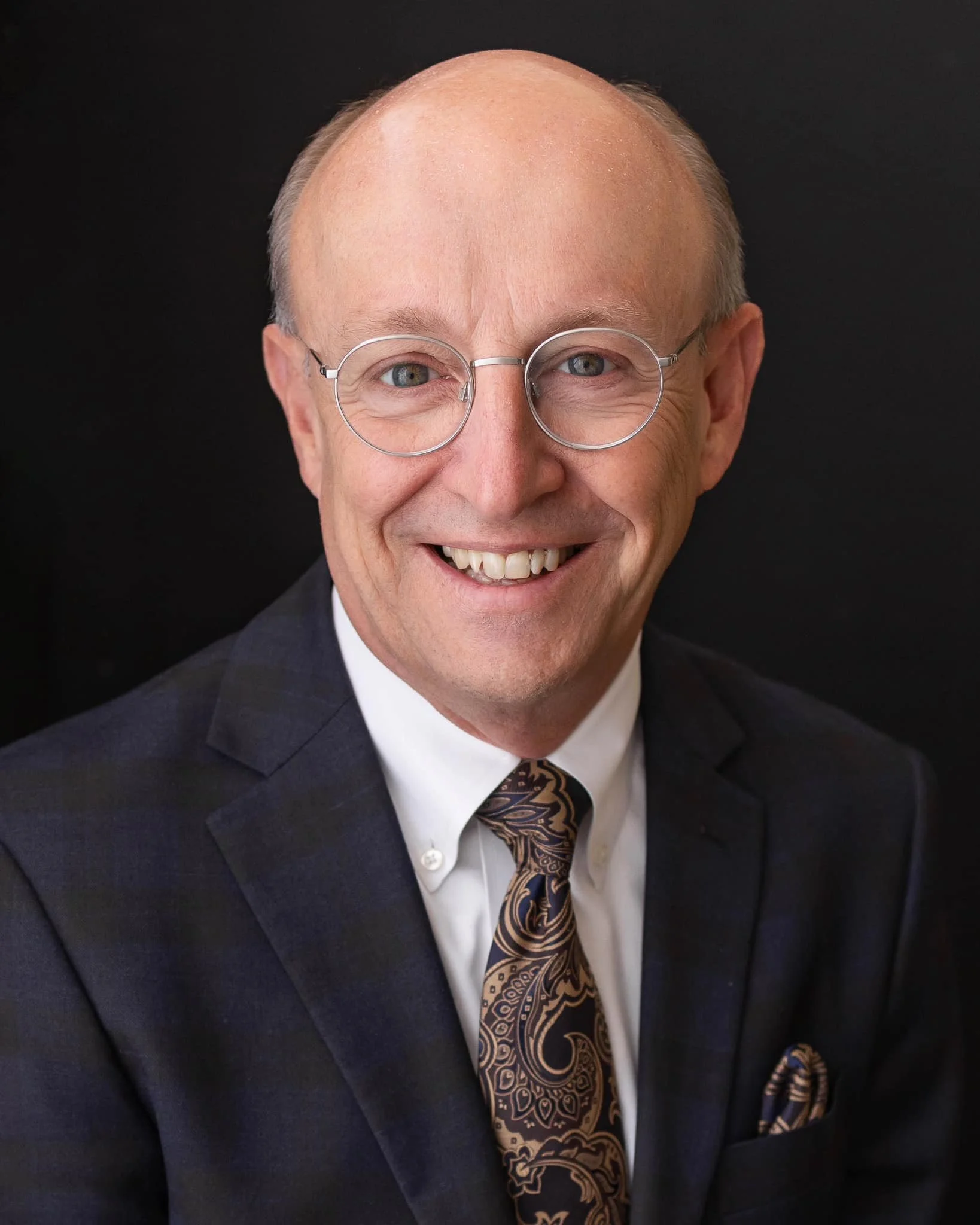Professional headshot of a smiling, bald man with glasses, wearing a dark plaid suit, white shirt, and a patterned tie against a black background.