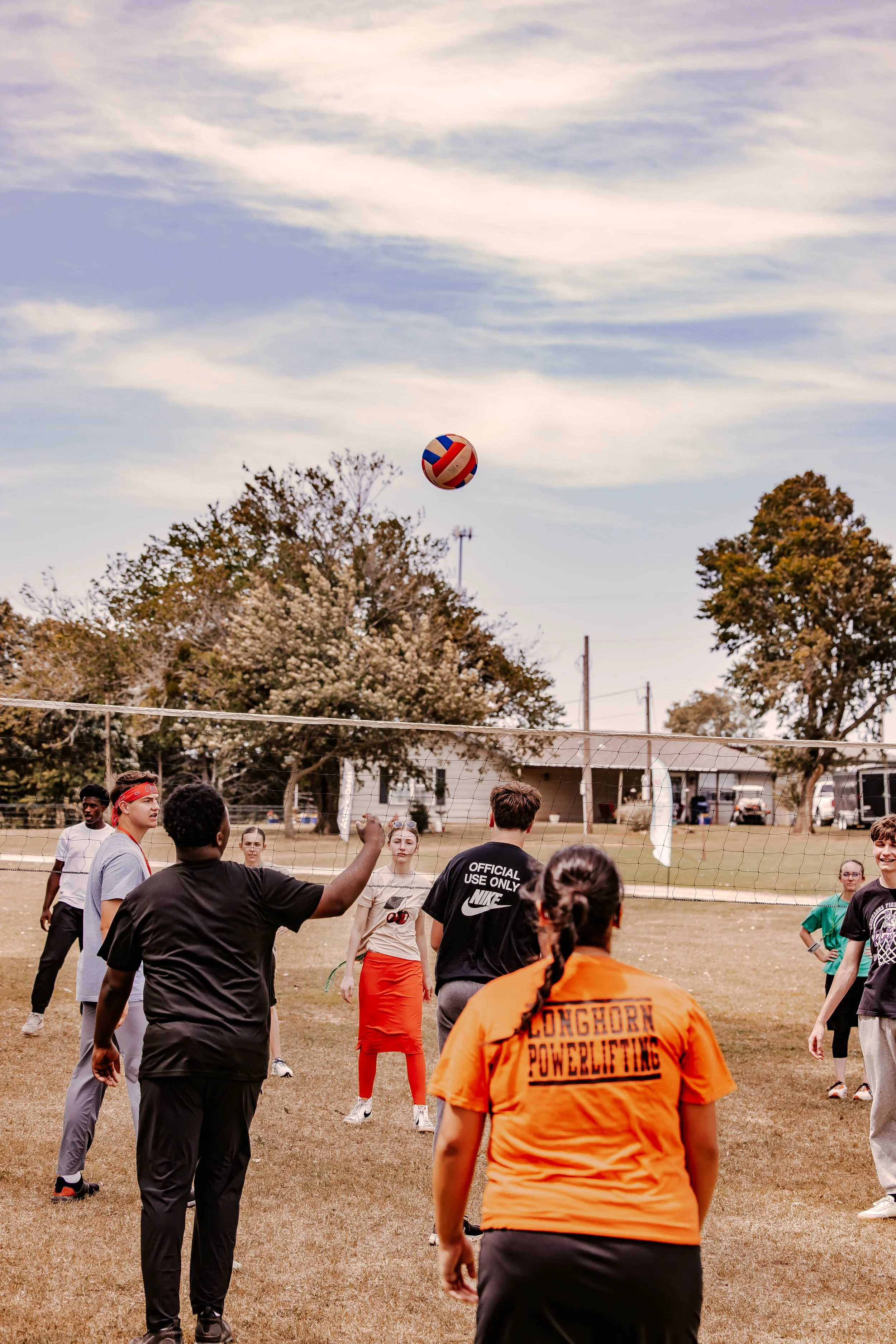 A group of people playing volleyball outdoors on a grassy field, with a volleyball in the air and trees in the background.