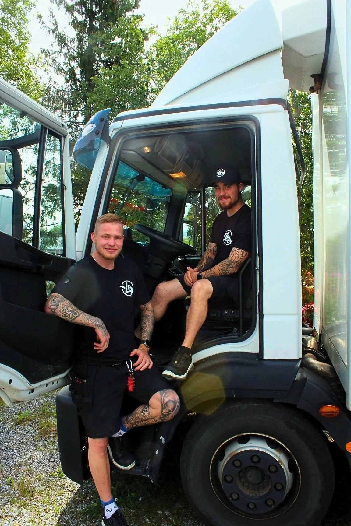 Logistiikkamestarit poseeravat kuorma-auton kanssa. Toinen istuu kuljettajan paikalla, ja toinen seisoo oven vieressä. Taustalla on puita ja selkeä taivasta.