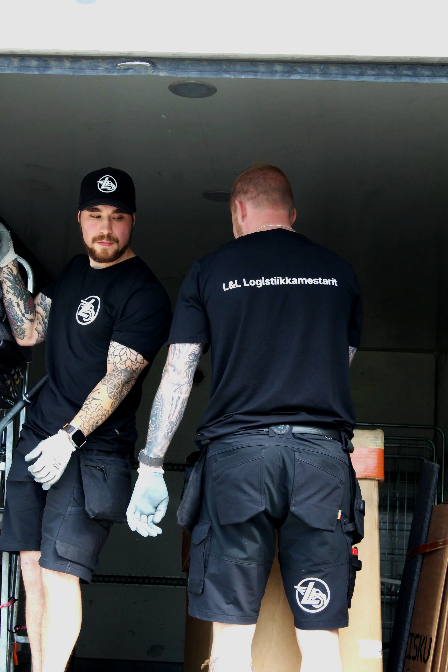 L&L Logistiikkamestarit siirtävät tavaroita kuorma-auton taka-osassa. Molemmilla yllään yrityksen logolla varustettu työasu.