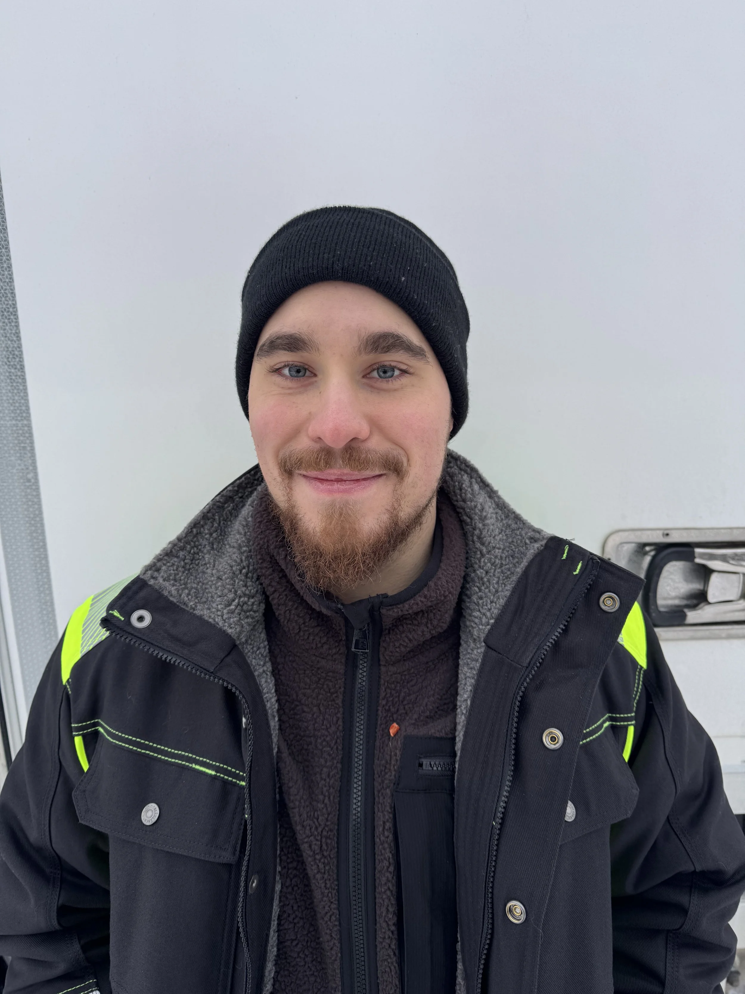 Henkilö hymyilee ja poseeraa kameralle. Hänellä on tummat hiukset ja parta, ja hän pukeutuu harmaaseen t-paitaan.