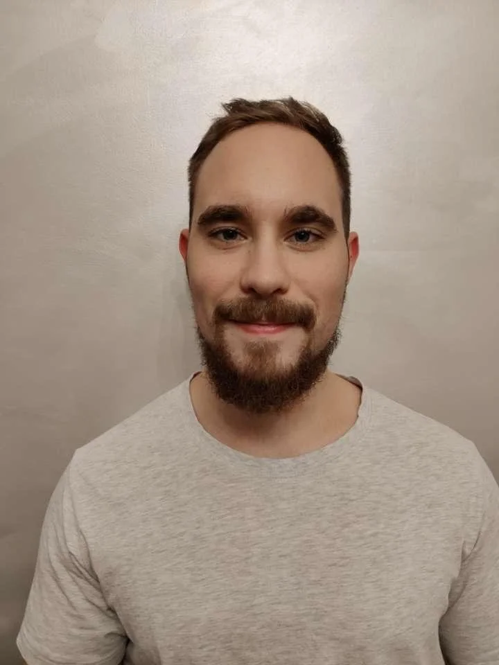 Henkilö hymyilee ja poseeraa kameralle. Hänellä on tummat hiukset ja parta, ja hän pukeutuu harmaaseen t-paitaan.