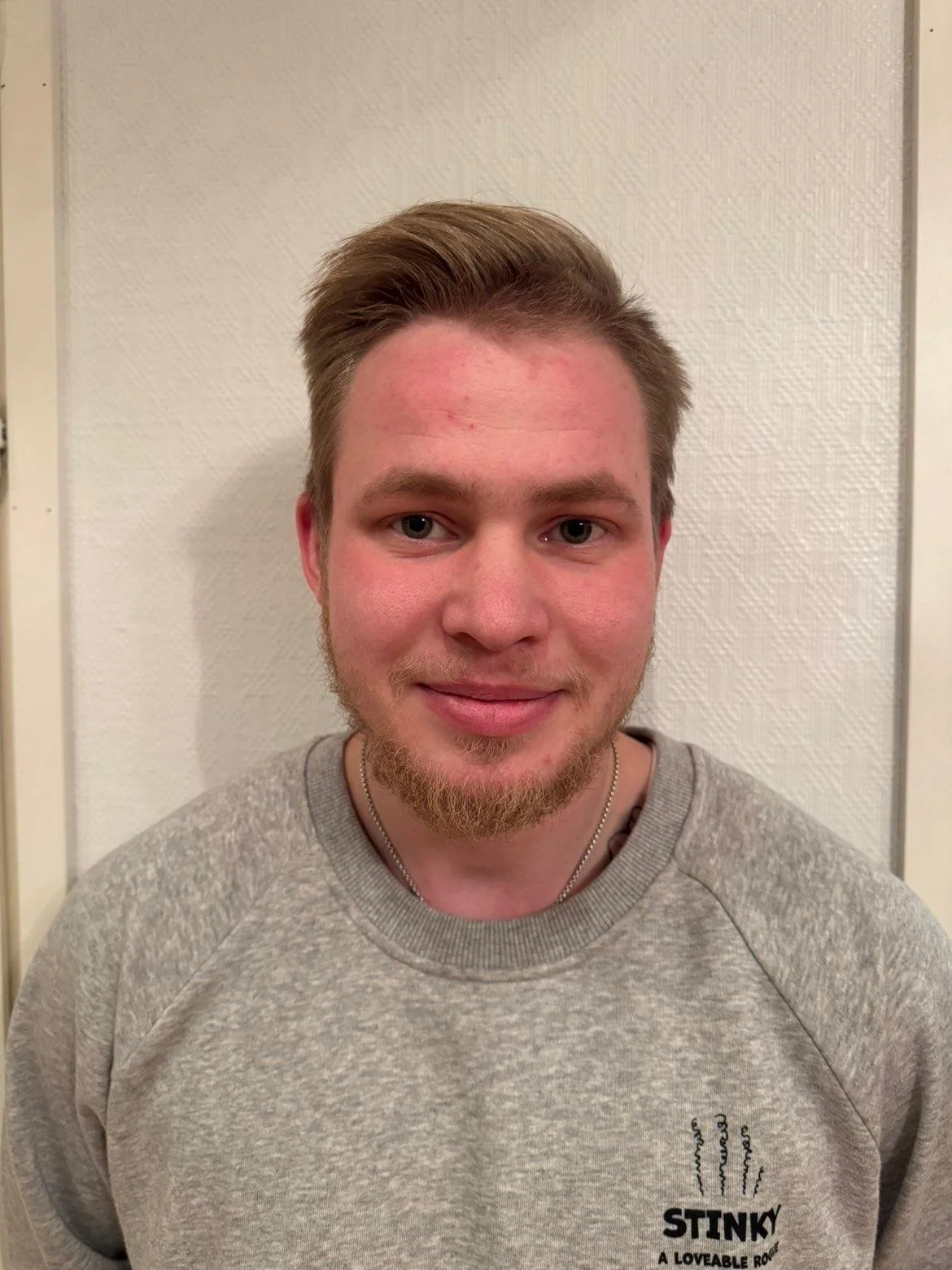 Nuori mies hymyilee, hänellä on lyhyet vaaleat hiukset, parta ja t-paita, jossa lukee 'STINKY A Loveable Roly'.