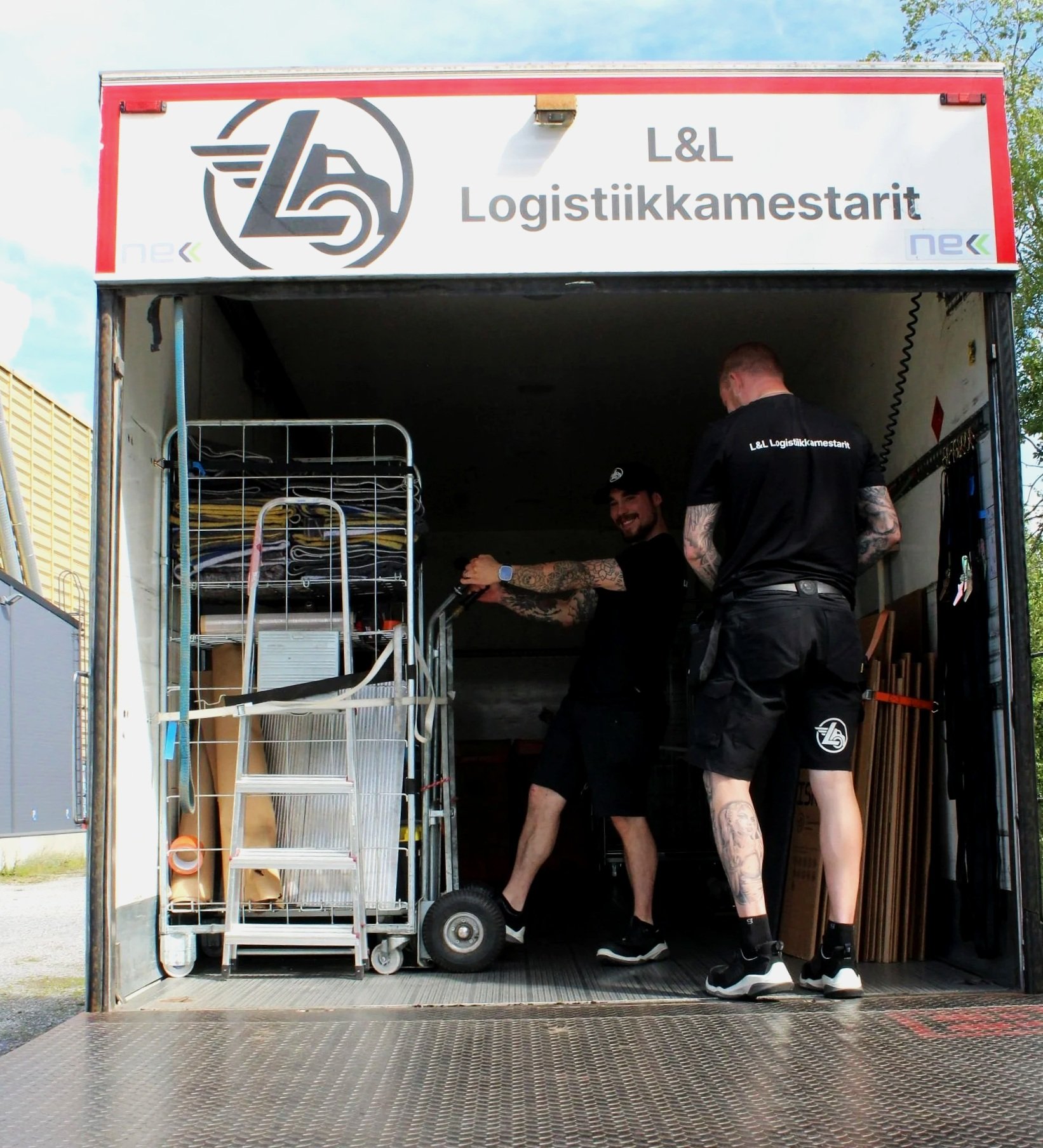 L&L Logistiikkamestarit toiminnassa, kaksi nuorta miestä lastaamassa tavaroita kuorma-autosta, yksi katsoo kameraan hymyillen.