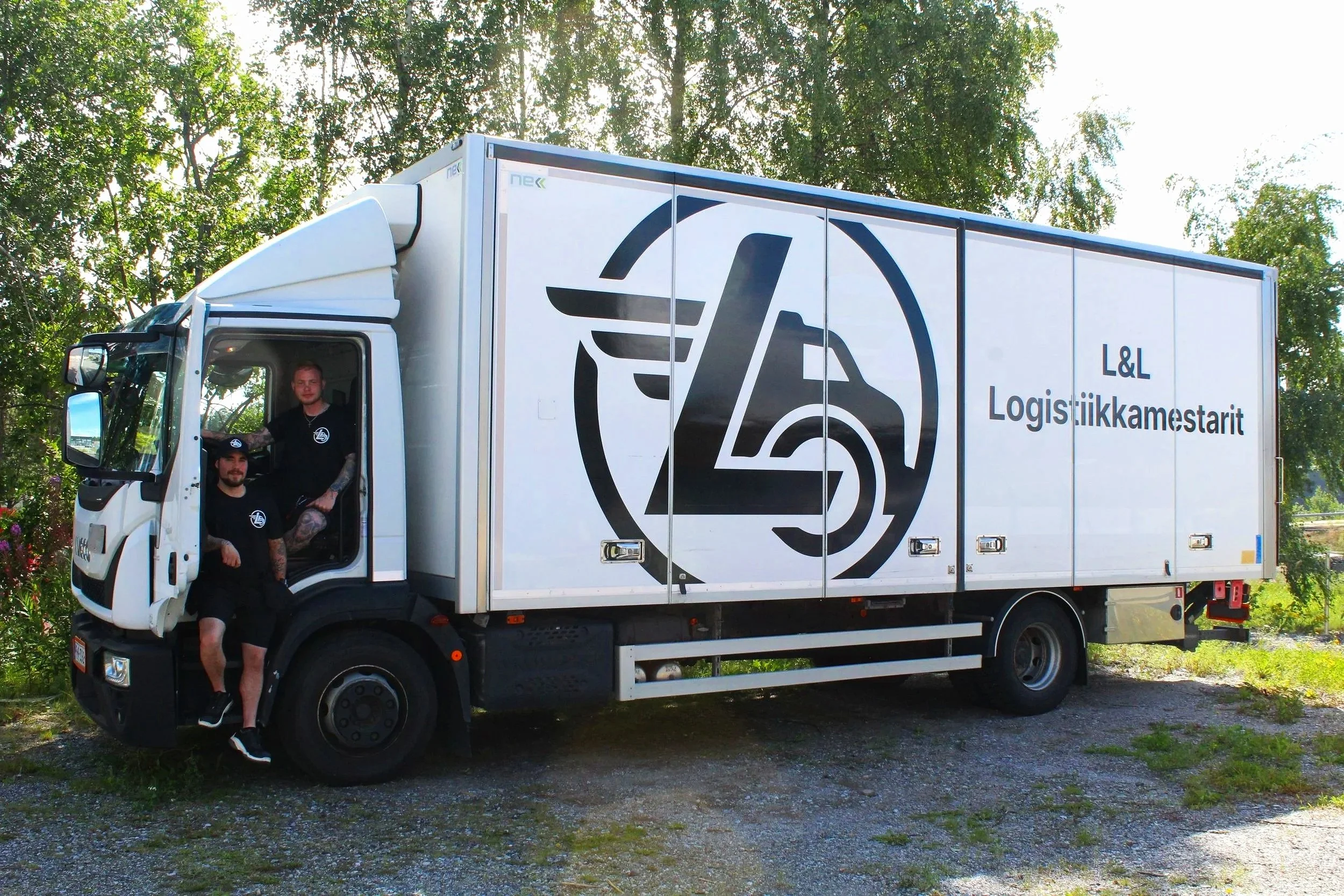Kuorma-auto, jossa on L&L Logistiikkamestarit-logo, ja kaksi miestä istuu sisällä. Taustalla puita ja sininen taivas.