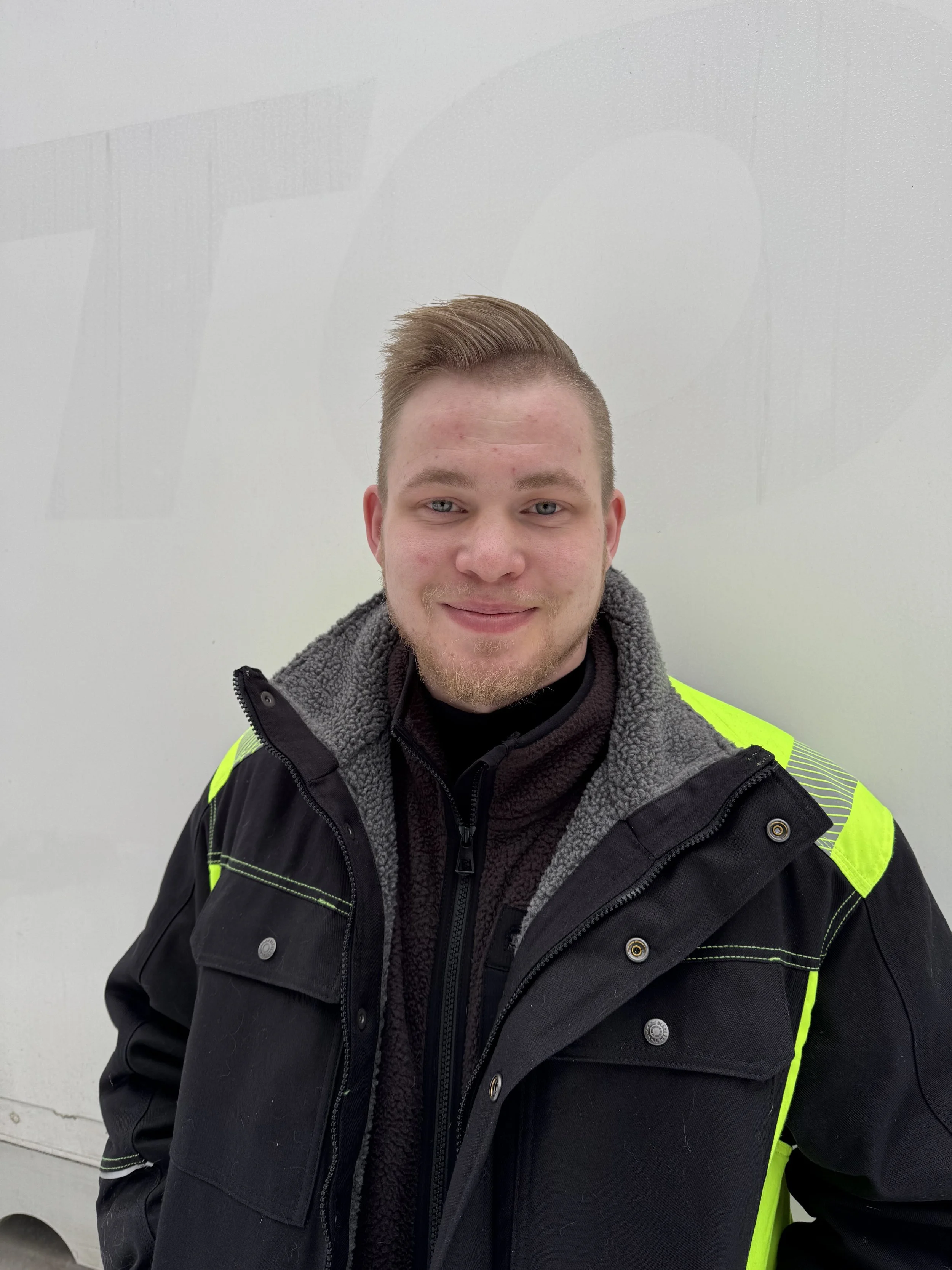 Nuori mies hymyilee, hänellä on lyhyet vaaleat hiukset, parta ja t-paita, jossa lukee 'STINKY A Loveable Roly'.