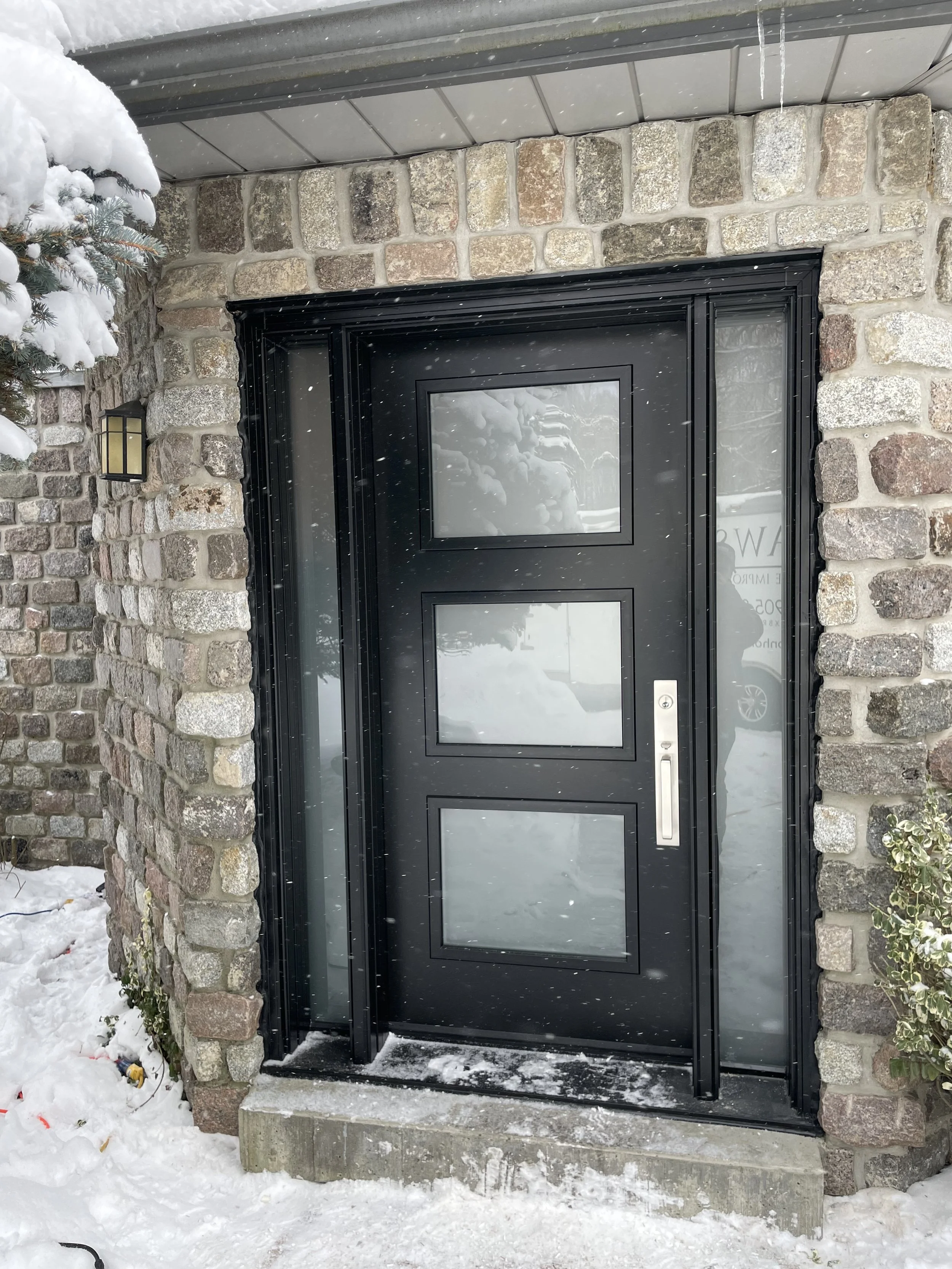 Black front door with three rectangular glass panels, surrounded by brick wall, with snow on the ground and snow falling.