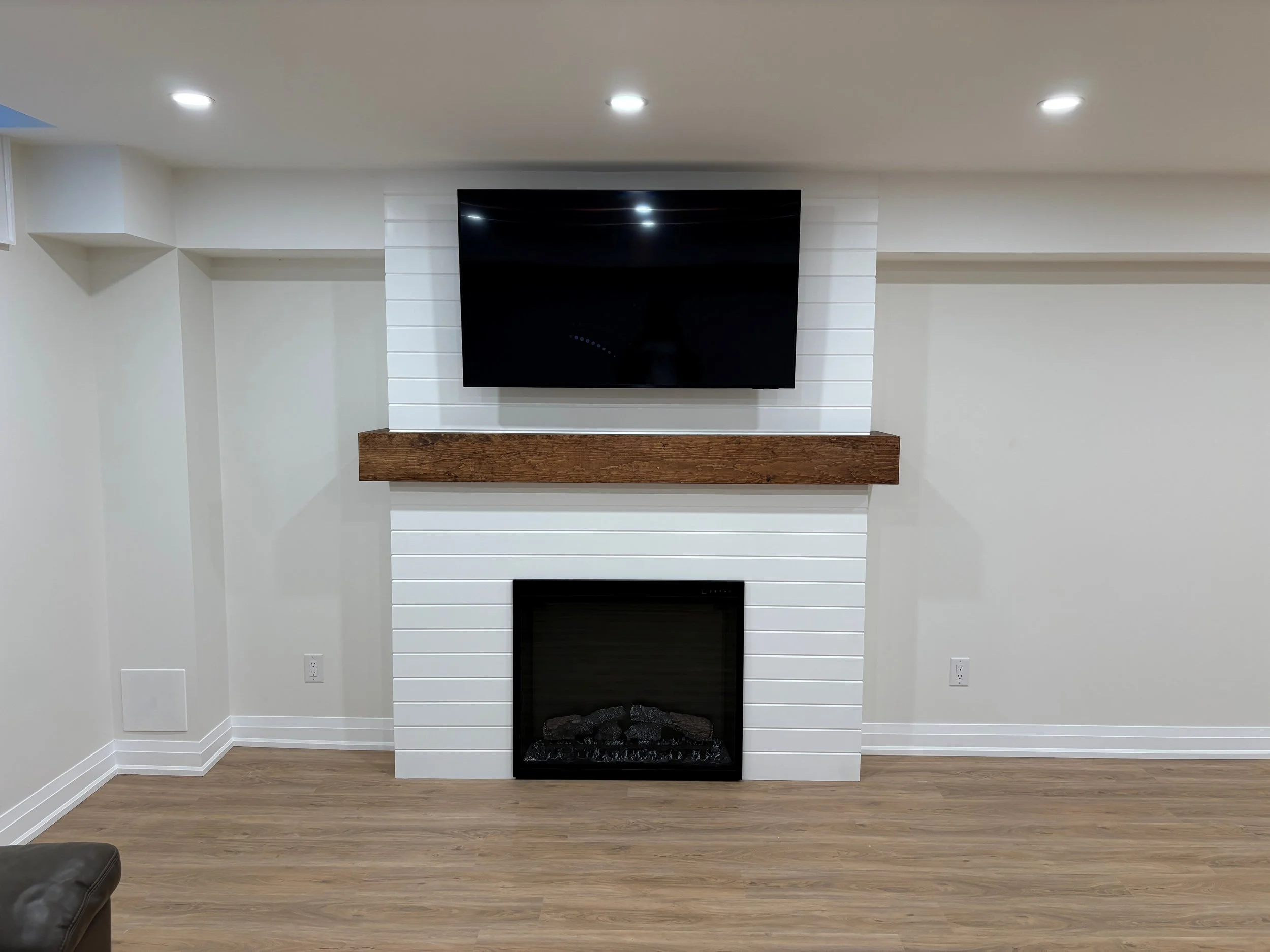 Living room with a fireplace and a mounted flat-screen TV above it. The fireplace has a black frame and stones inside, and a wooden mantel above. The walls and ceiling are painted white, with recessed lighting. The floor has light wood flooring.