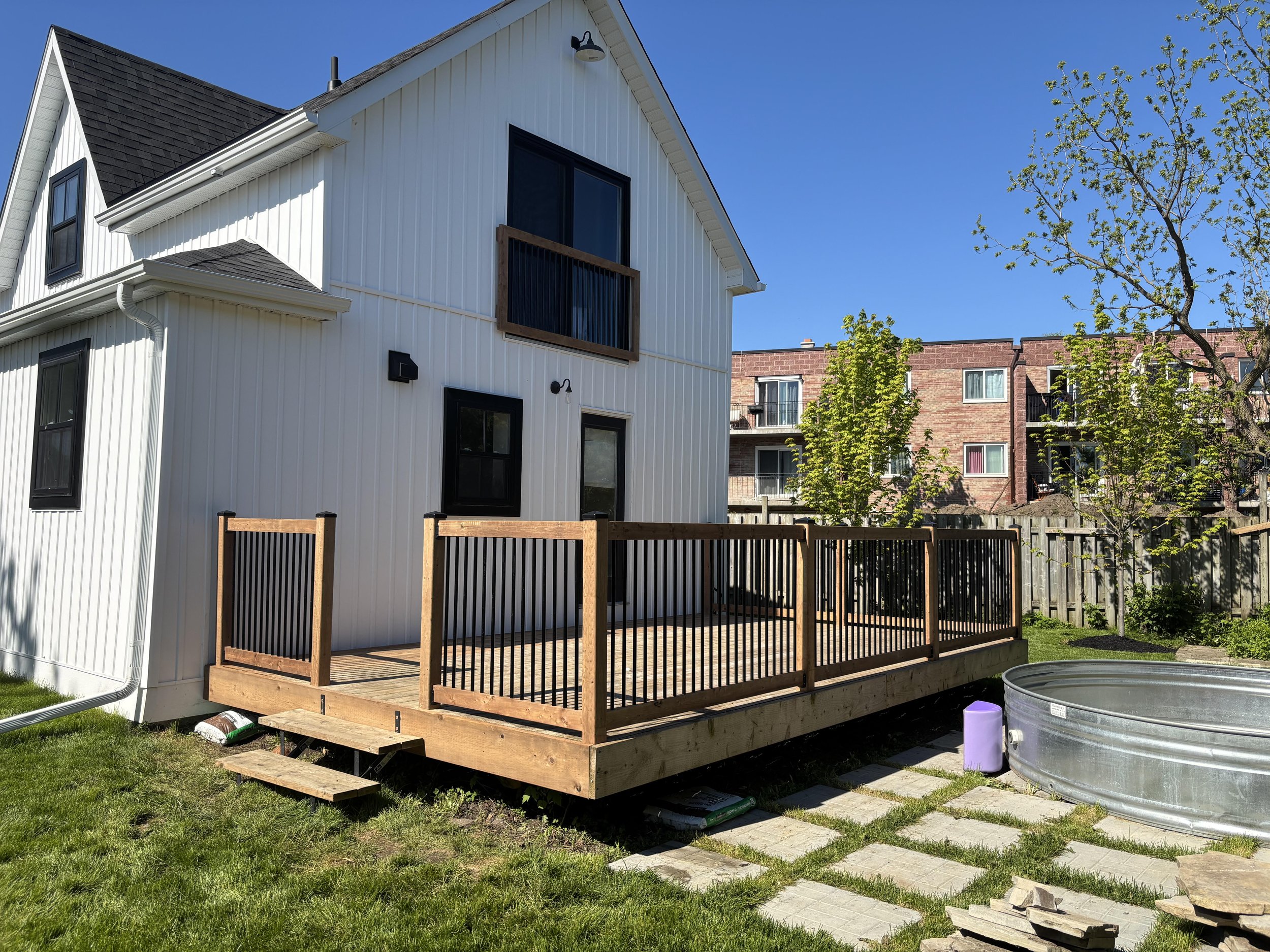 Backyard with a newly built wooden deck, attached to a white house with black-trimmed windows, and a small above-ground metal pool. There are trees, a grassy lawn, and neighboring multi-story brick apartment buildings.