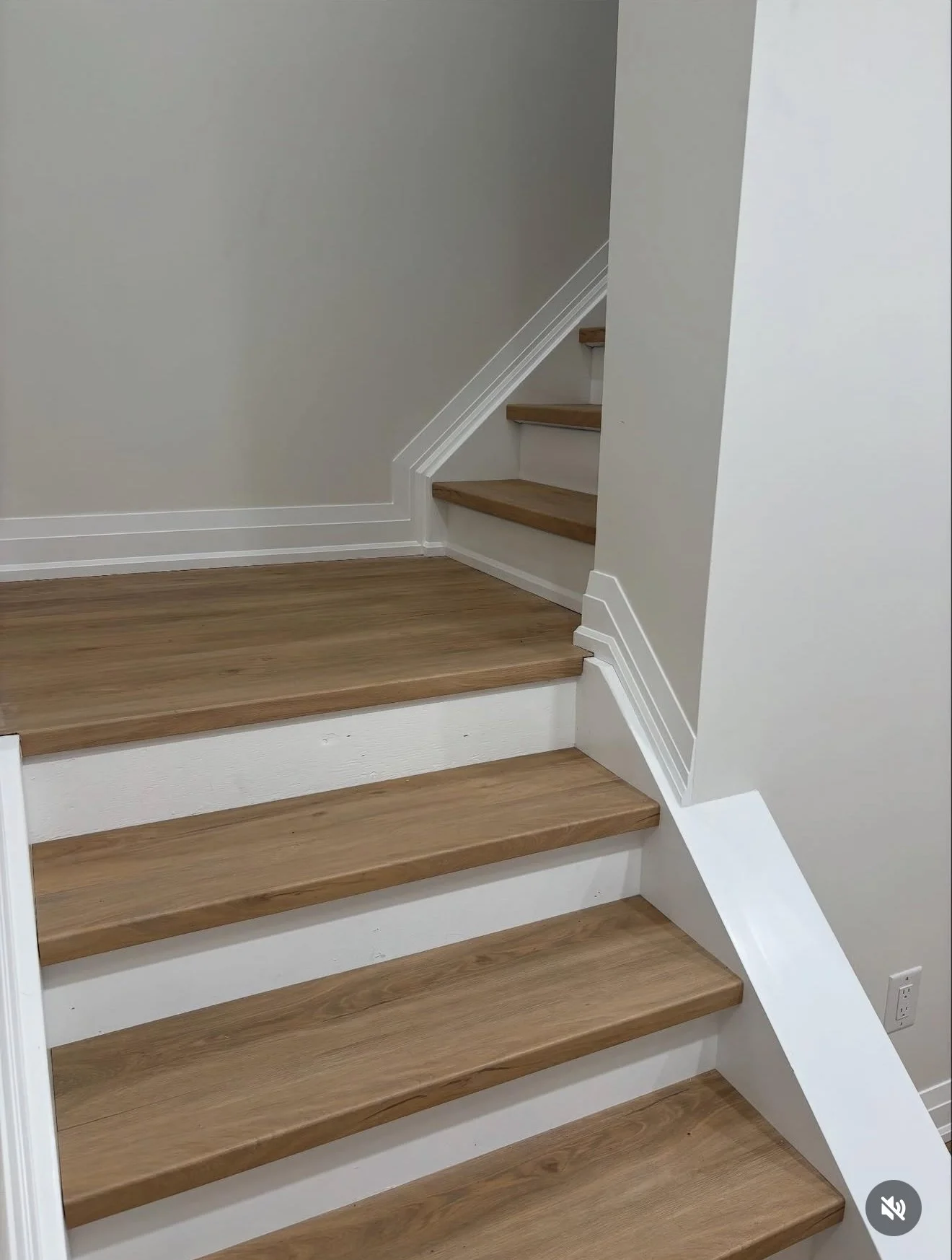 Wooden stairs with white risers, leading up to a small landing with a short staircase to the right, against white walls.