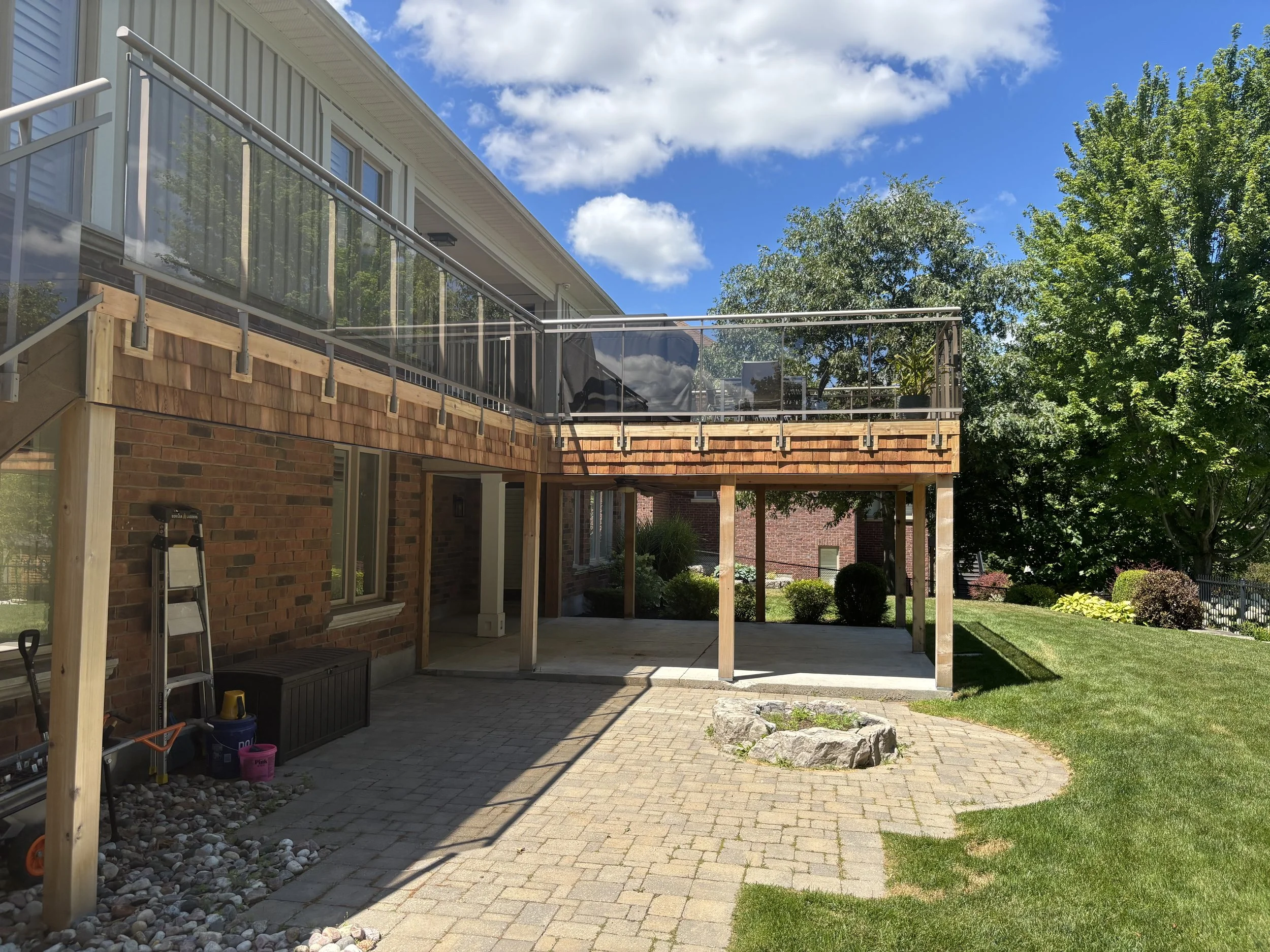 Backyard with wooden deck, brick house, paver patio, grassy area, trees, and a cloudy sky.