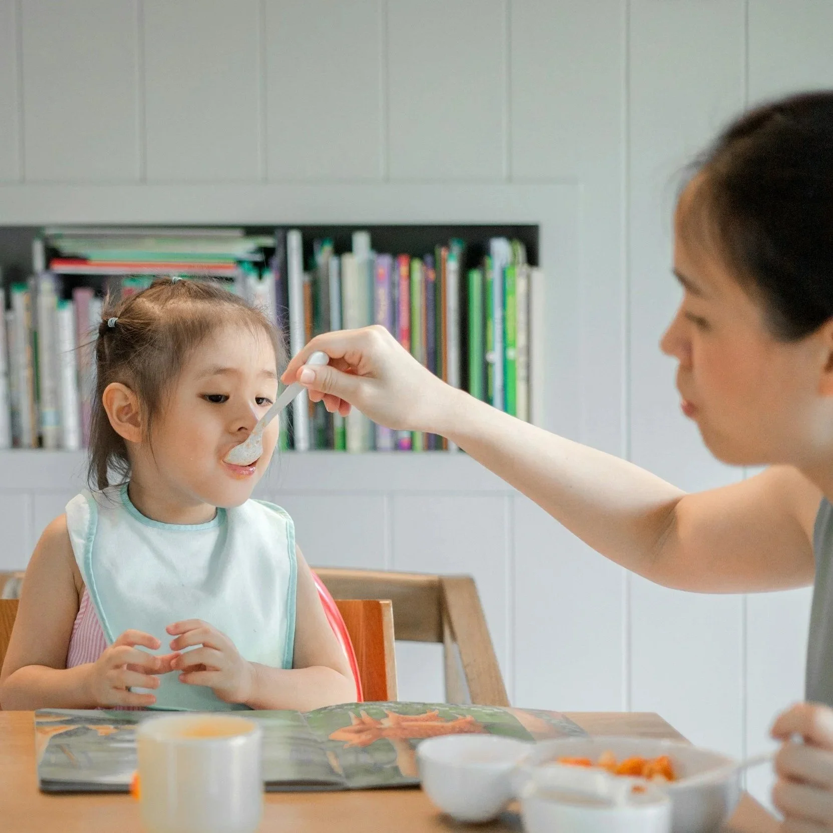 A woman is feeding a young girl with a spoon in a room with bookshelves in the background. The girl is wearing a bib and sitting at a table with bowls of food and cups.