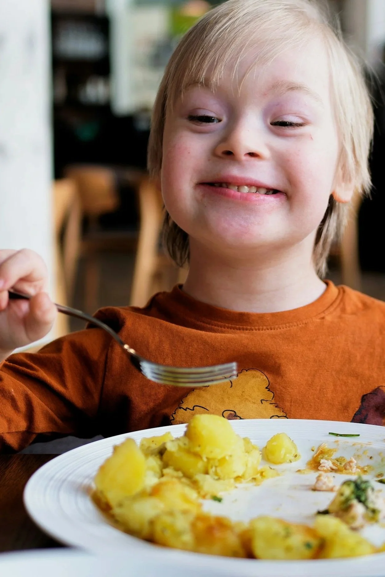 A young boy with blond hair smiling while holding a fork over a plate of food, with a blurred background of a dining area.
