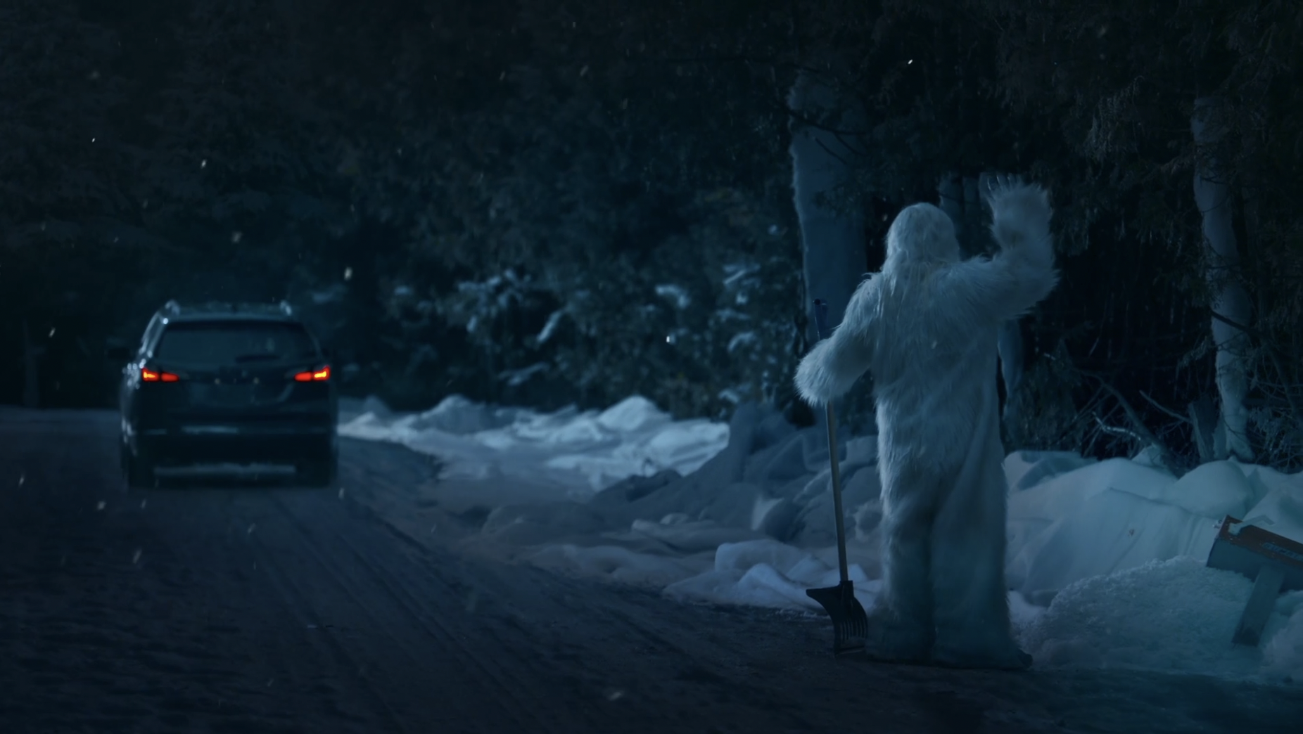A person dressed in a white, furry costume resembling a yeti or Bigfoot, standing on a snowy roadside at night, holding a shovel, with a car approaching in the background.
