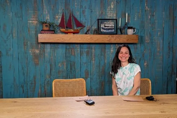 A woman with long dark hair smiling while sitting at a wooden table in front of a blue wooden wall with nautical decorations, including a model sailboat, framed pictures, and various small items on a floating shelf.