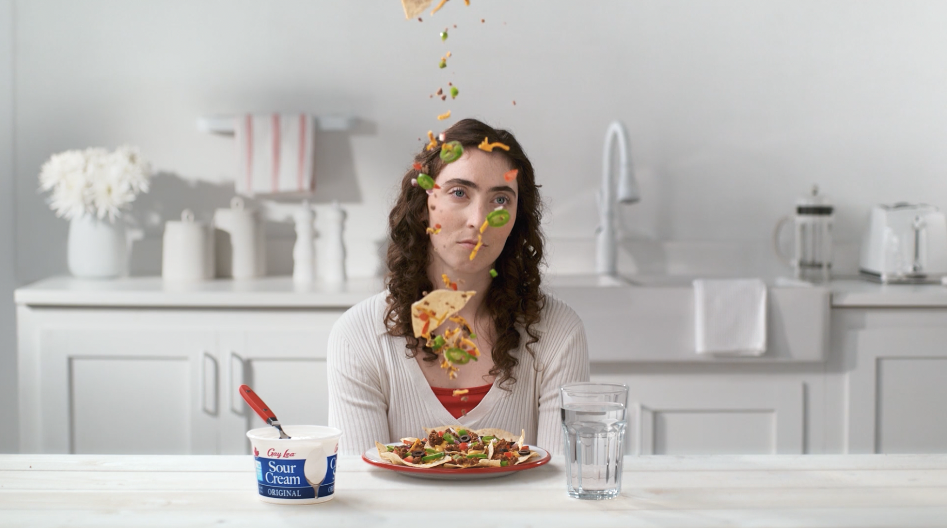 A woman has a slice of pizza thrown at her, with toppings spilling and flying through the air in a kitchen setting.