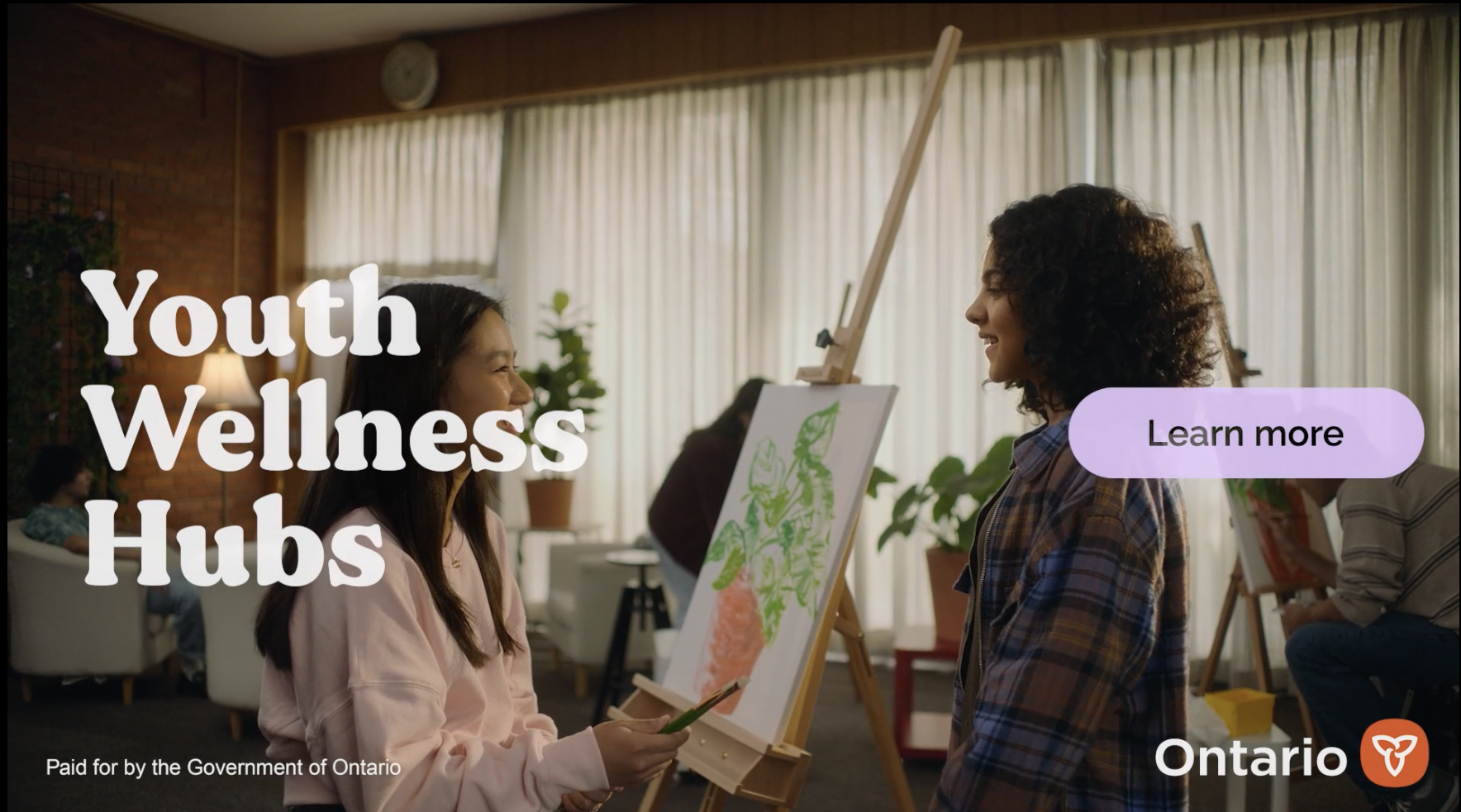 Two young women smiling and talking at an indoor youth wellness hub, with art supplies and painting easels around them, in a well-lit room with large curtains.