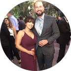 A man and a woman dressed formally at an outdoor event. The woman is wearing a maroon dress, and the man is in a gray suit with a tie. They are smiling and standing close together.