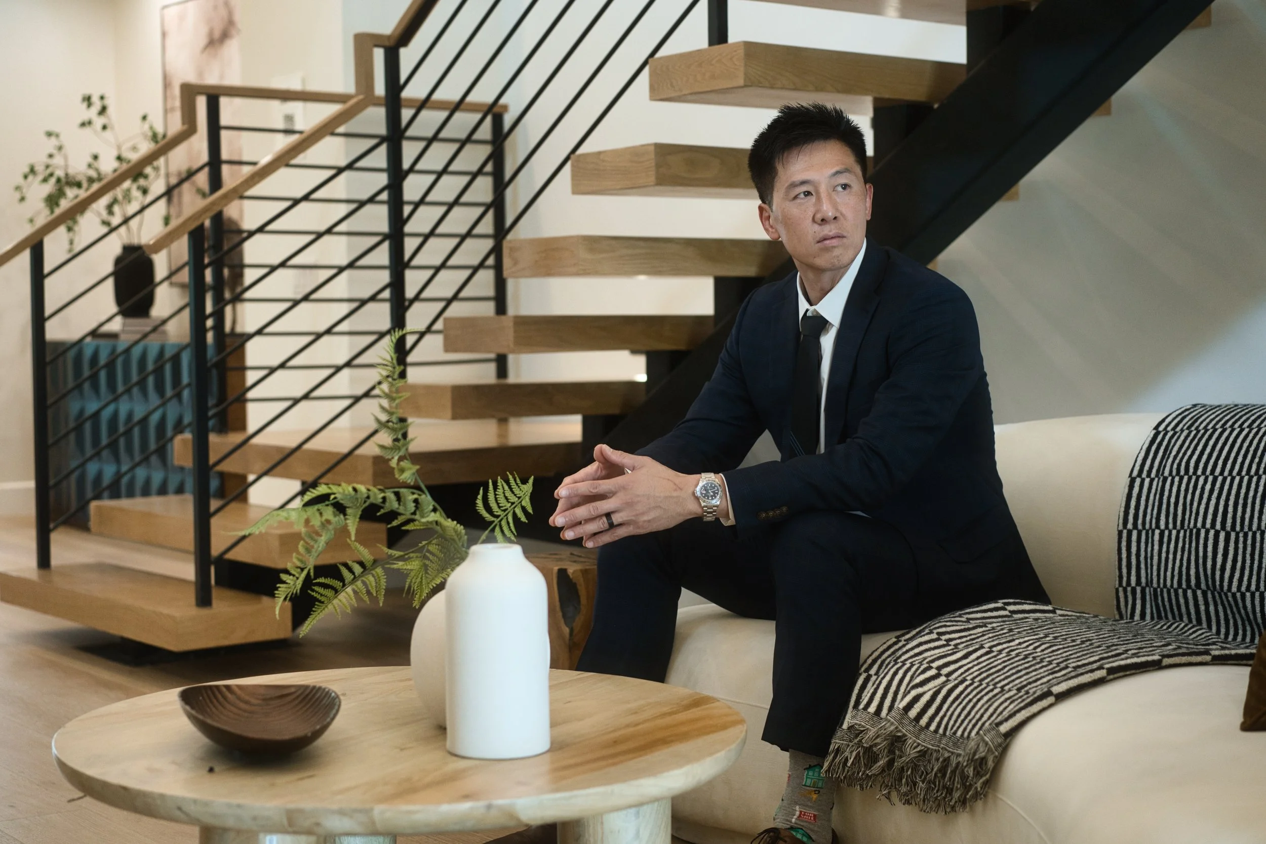 A man in a dark suit sitting on a cream-colored sofa in a modern living room, with a wooden staircase and minimalist decor including a white vase and a small bowl on a round wooden coffee table.