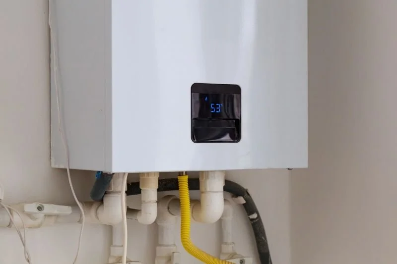 A white tankless water heater mounted on a wall with visible pipes and hoses below it, including yellow and black hoses, and a digital display showing the temperature.