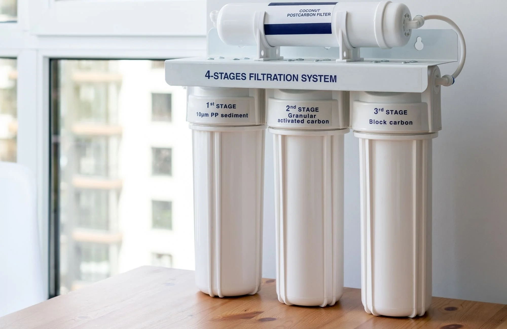 Four-stage water filtration system with labeled filters including 10um PP sediment, granular activated carbon, and block carbon on a wooden table near a window.
