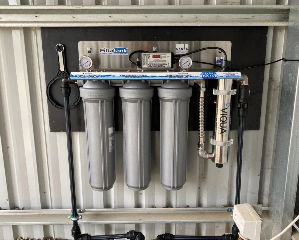 A water filtration system installed on a wall, featuring three large silver filter housings, a UV sterilizer labeled 'VIOUX,' gauges, and various pipes and fittings for water treatment.