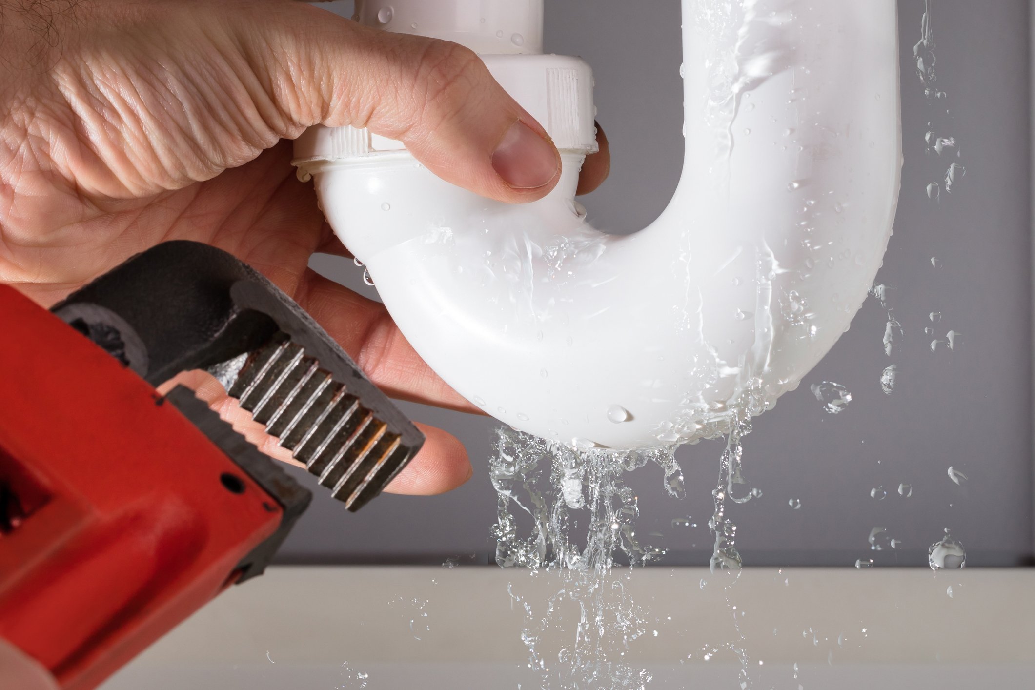 A hand holding a white plastic pipe under running water with a red and black pipe wrench nearby.