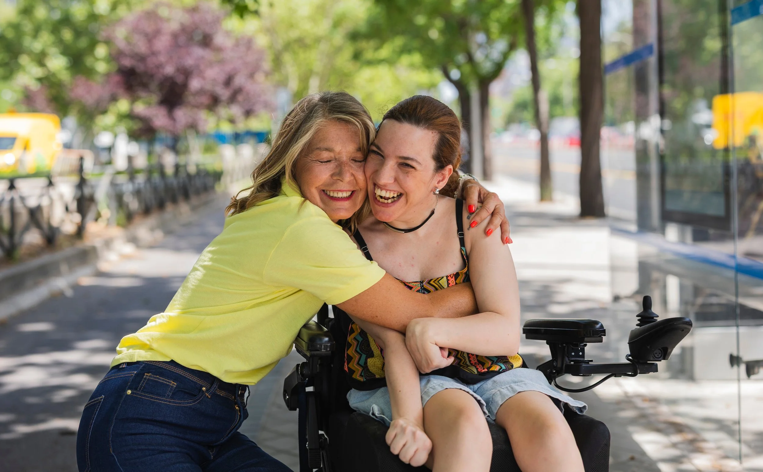 Two women smiling and hugging on a city sidewalk with trees and buses in the background.