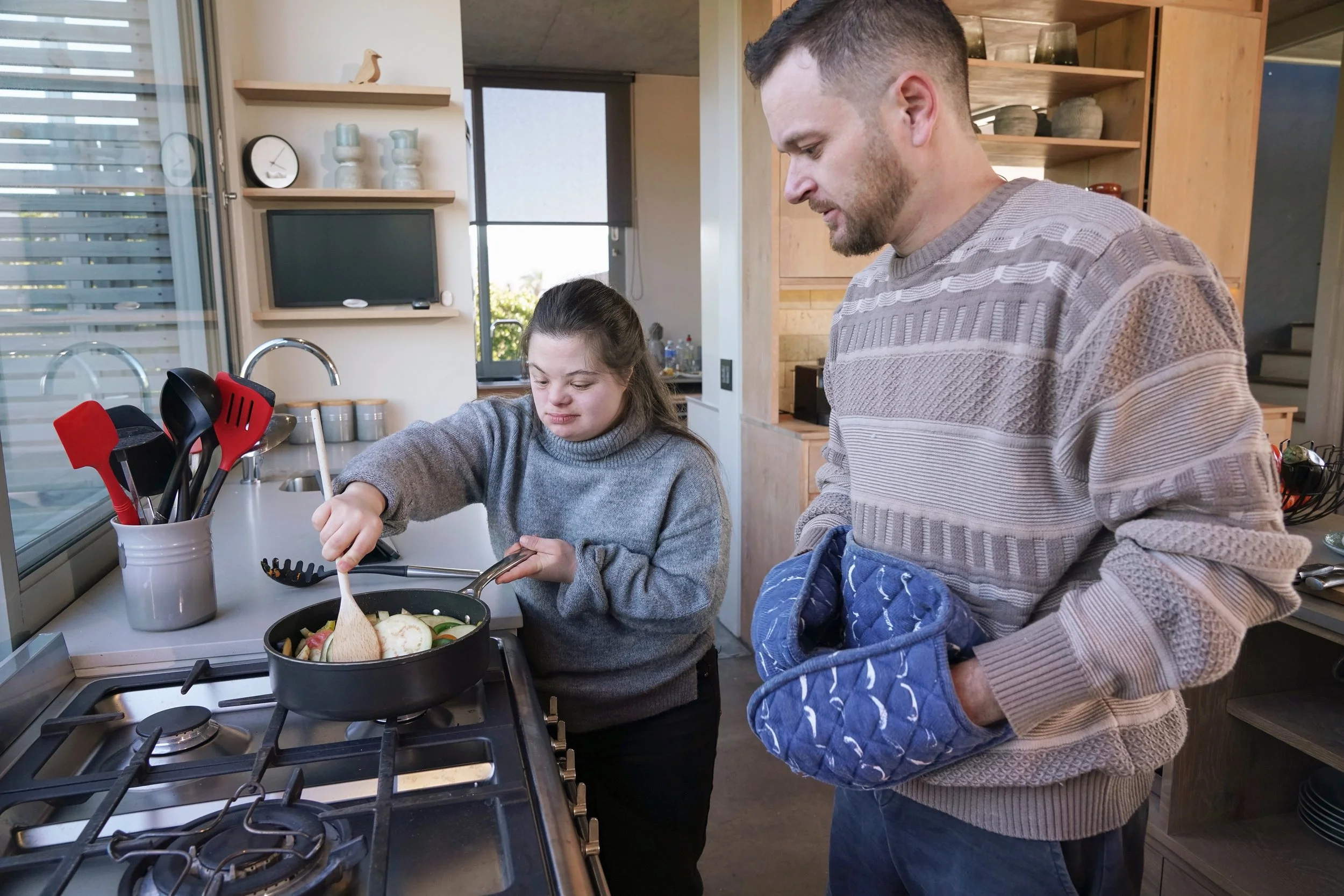 A man and a girl cooking together in a kitchen. The girl is stirring a pan on the stove, and the man is watching. The girl is wearing a gray sweater, and the man is wearing a patterned beige sweater and holding an oven mitt.