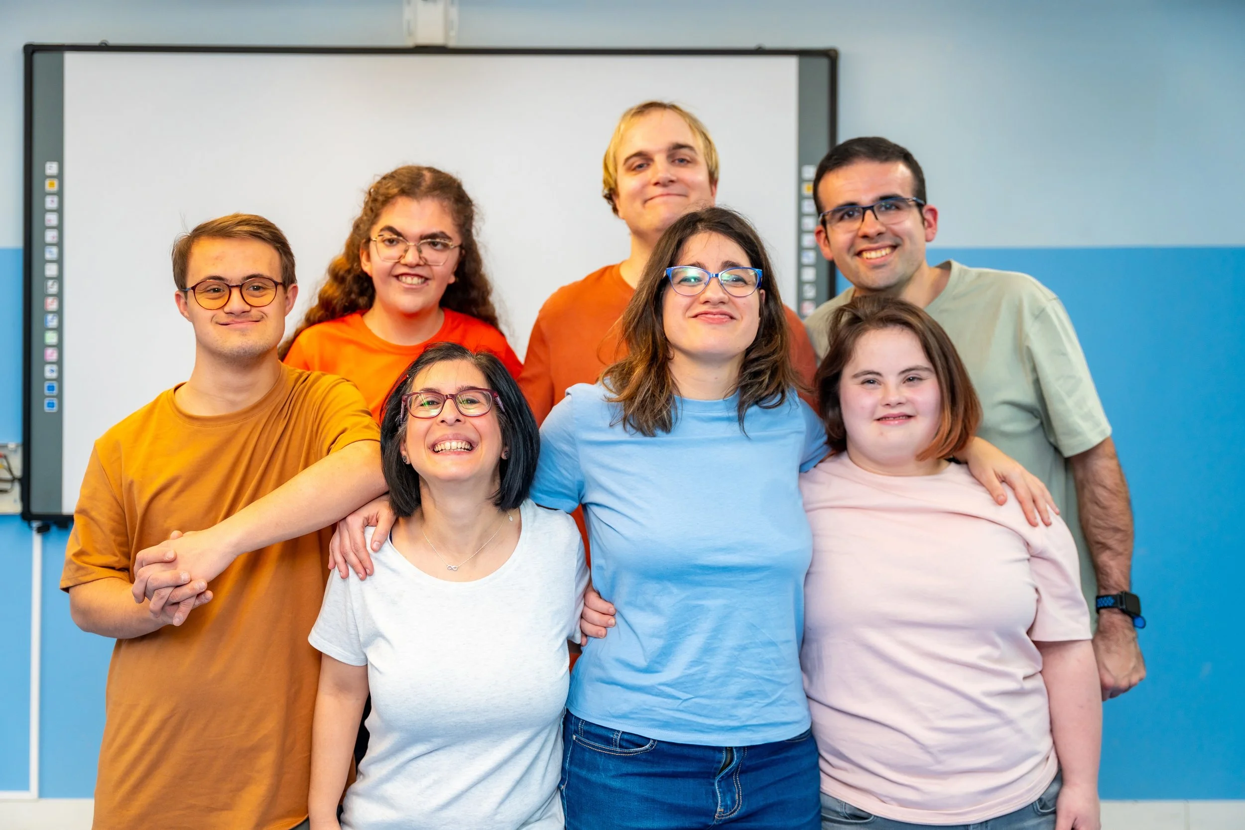 Group of eight diverse students smiling and posing together in a classroom, standing in front of a whiteboard.