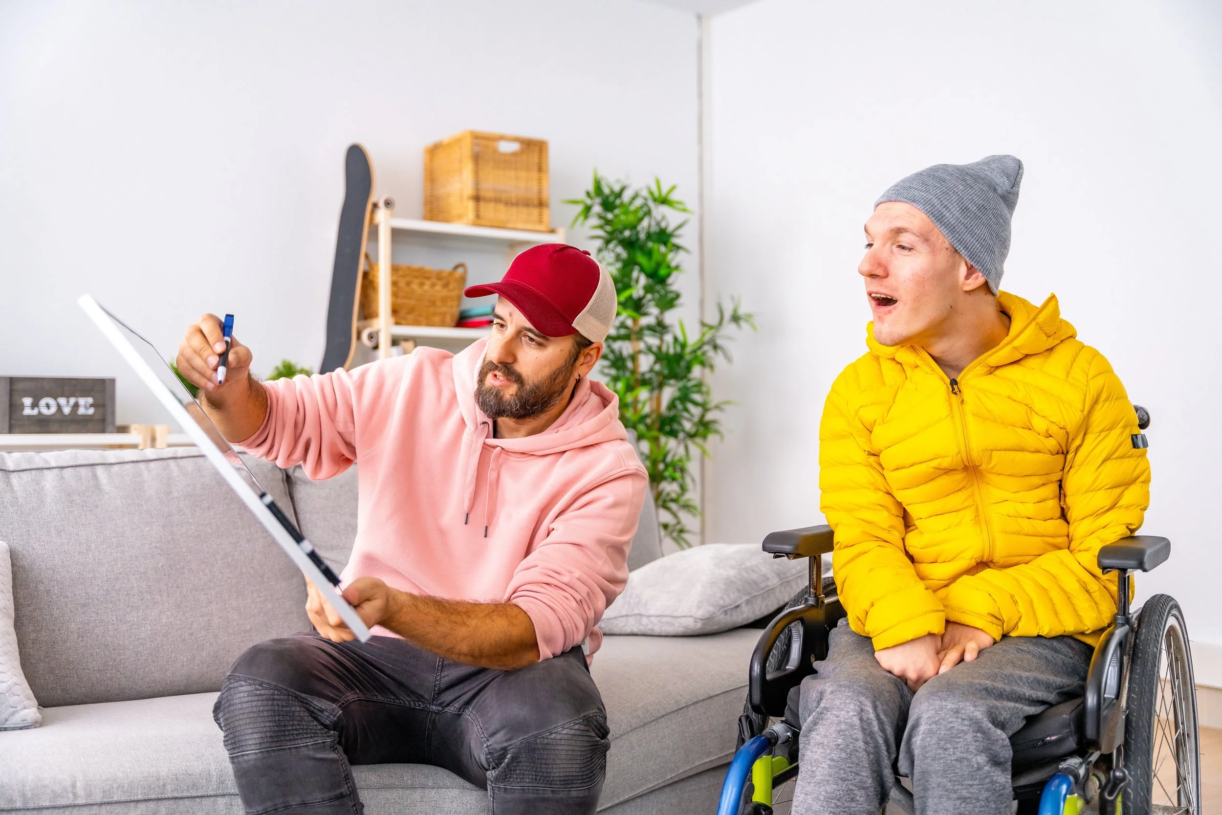 A man in a pink hoodie and red cap sitting on a couch, showing something on a whiteboard to a young man in a wheelchair wearing a yellow jacket and gray beanie, in a cozy living room with a skateboard, shelving, and houseplants in the background.
