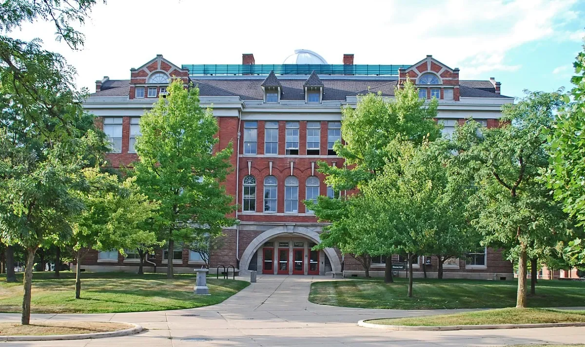 A large brick building with arched windows and a central entrance, surrounded by green trees and a grassy lawn.