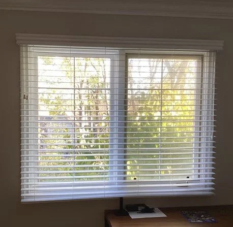 A window with white blinds partially open, showing trees and greenery outside.