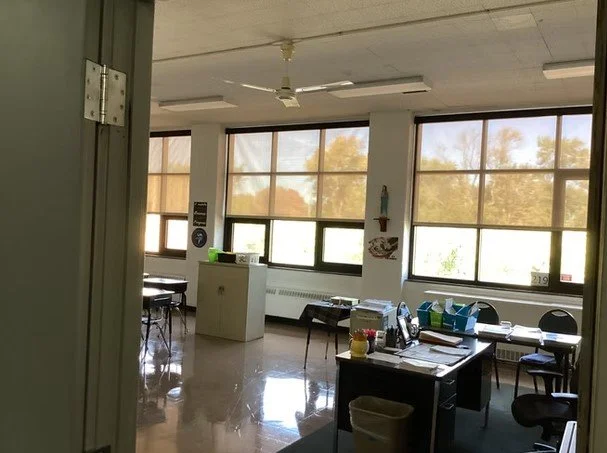 An empty classroom with large windows, desks, chairs, and a teacher's desk with office supplies.