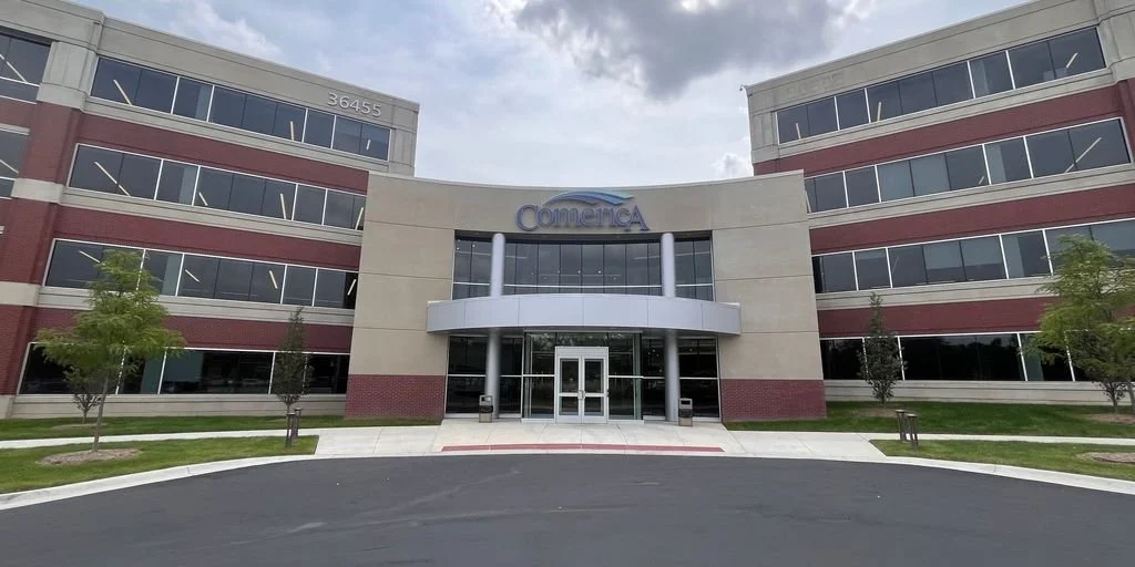 Front view of a modern corporate office building with the sign 'Comerica' above the entrance, surrounded by a driveway and landscaping with small trees.