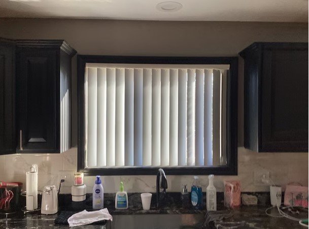 Kitchen window with vertical blinds, dark cabinets, and various items on the countertop including a paper towel roll, soap dispenser, and small appliances.