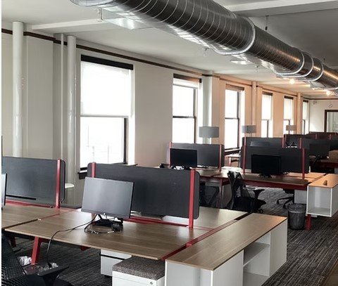 Empty office with rows of desks, computer monitors, and large windows.