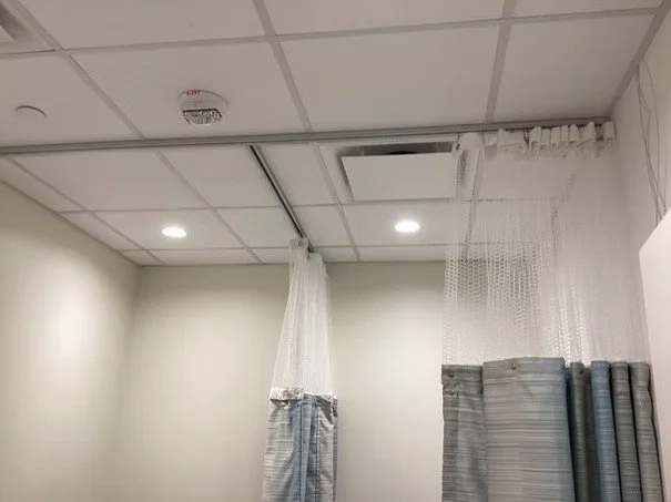 Ceiling with a drop ceiling grid, two ceiling lights, a partially open ceiling vent, and hospital curtains hanging from the ceiling.