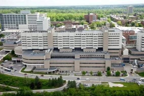 A large multi-story hospital building with a parking lot and several cars, surrounded by trees and cityscape in the background.