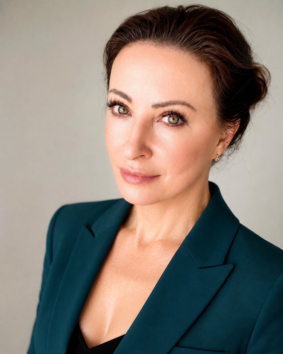 Close-up portrait of a woman with short brown hair, green eyes, and light skin, wearing a dark teal blazer over a black top, smiling slightly against a light grey background.