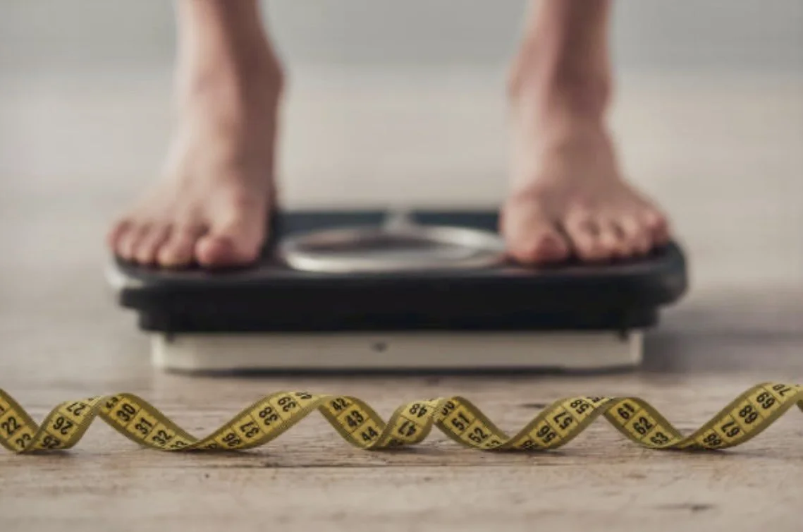 A person standing on a digital weighing scale, with a yellow measuring tape in the foreground on a wooden floor.