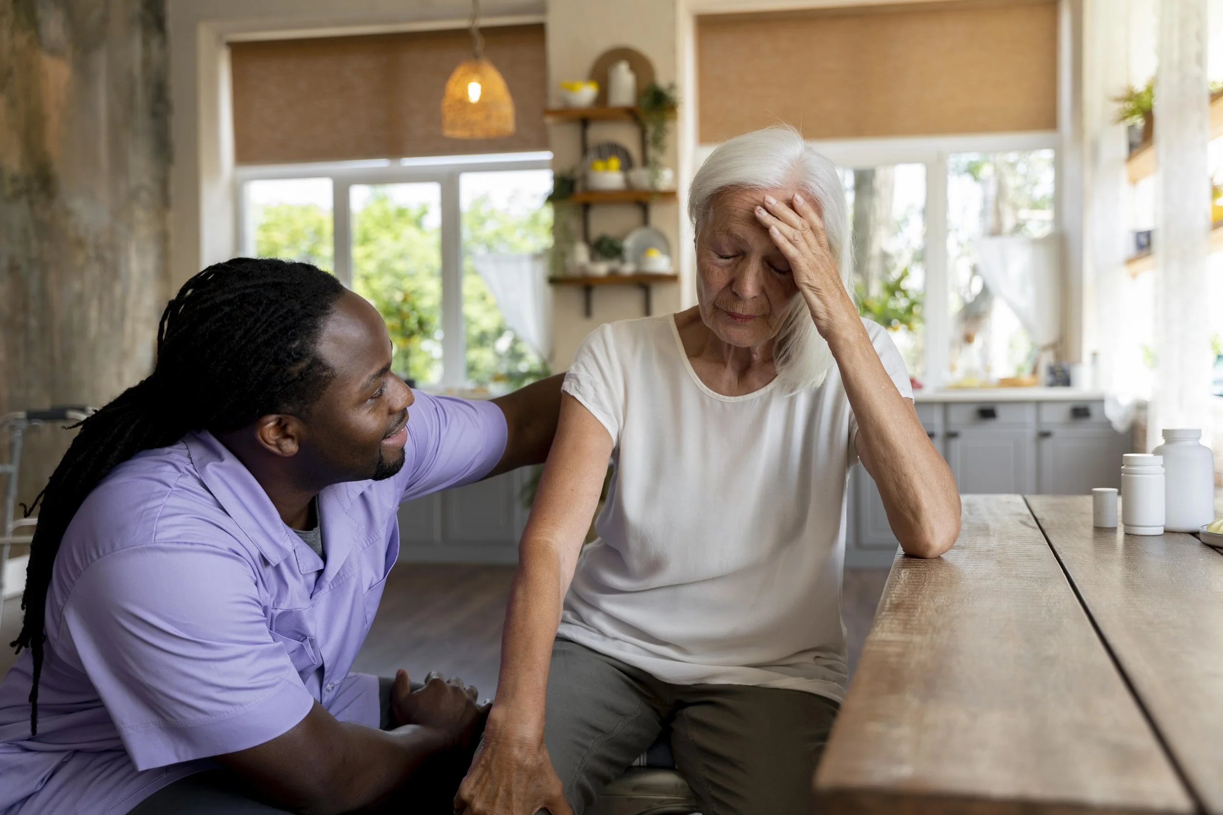 A young caregiver with dreadlocks and purple scrubs comforting an elderly woman with white hair, who looks distressed and is sitting at a wooden table in a bright, sunny kitchen.