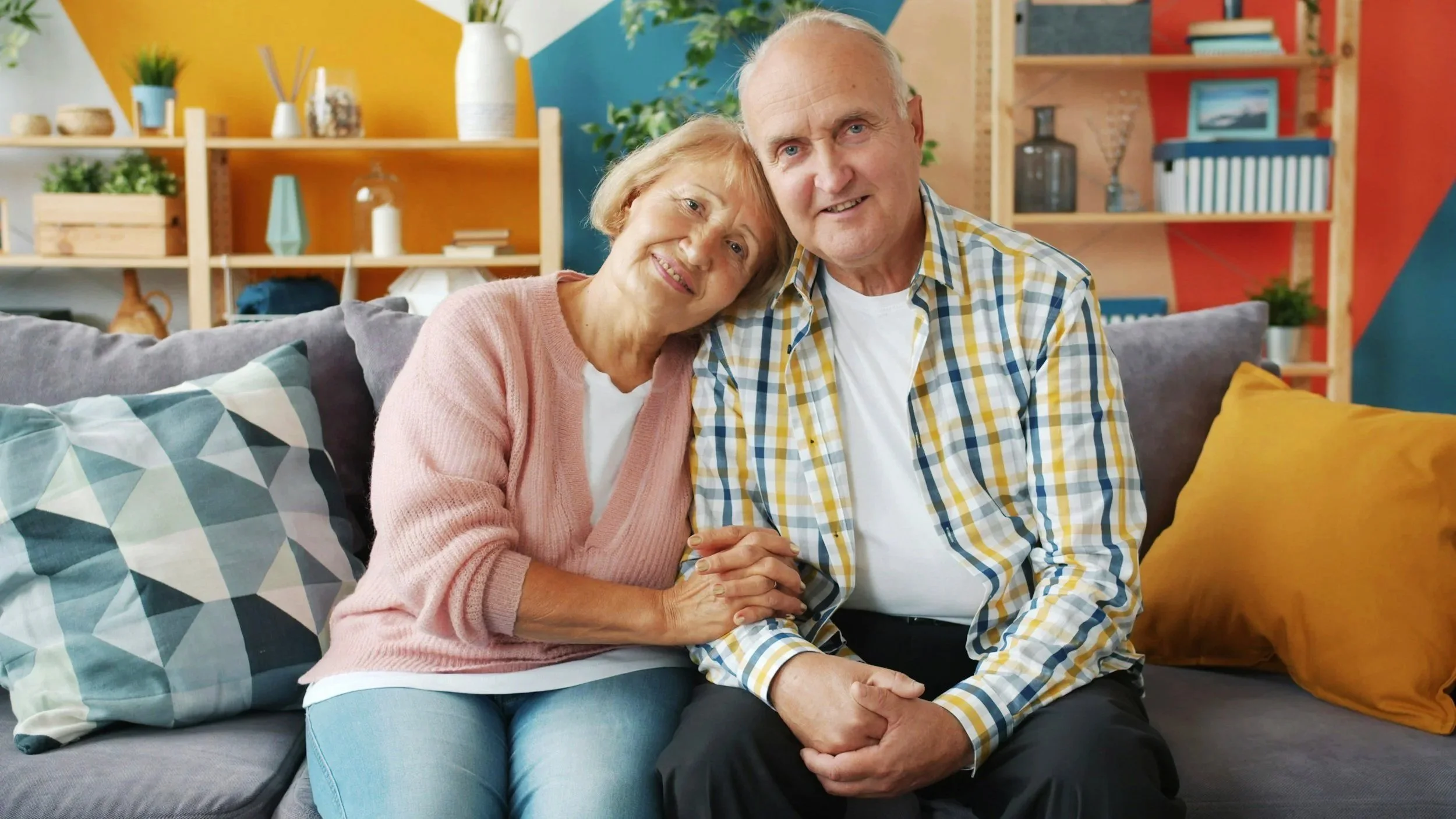 An elderly couple sitting close together on a colorful sofa in a cozy living room, smiling and holding hands.