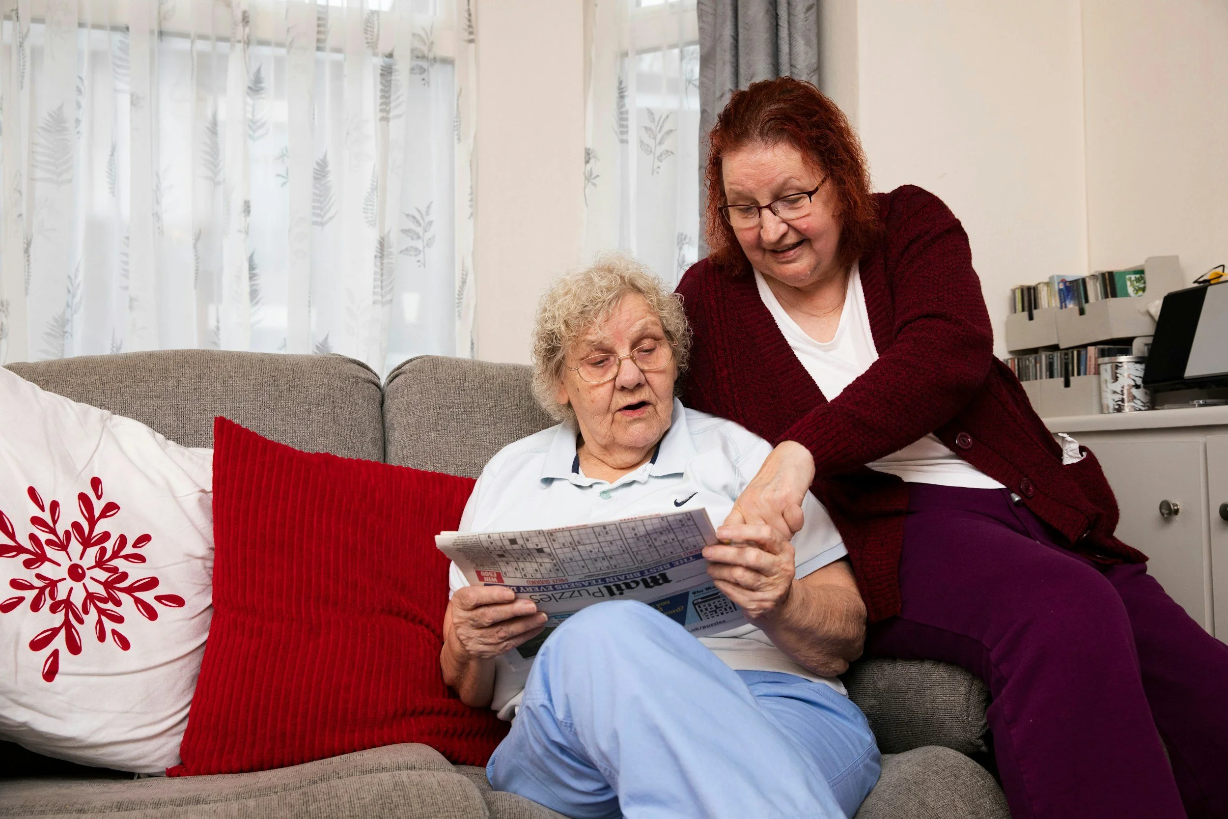 An elderly woman sitting on a grey couch, reading a newspaper with a younger woman leaning over her, pointing at the newspaper, in a living room with white curtains and bookshelves.