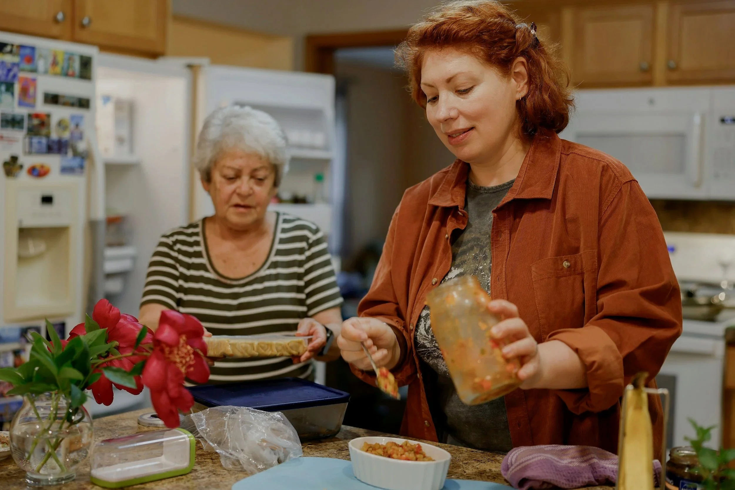 Two women preparing food in a kitchen. One woman with short, gray hair and a striped shirt is holding a tray, while the other woman with curly, red hair and an orange shirt is spooning food from a jar into a bowl on the countertop.