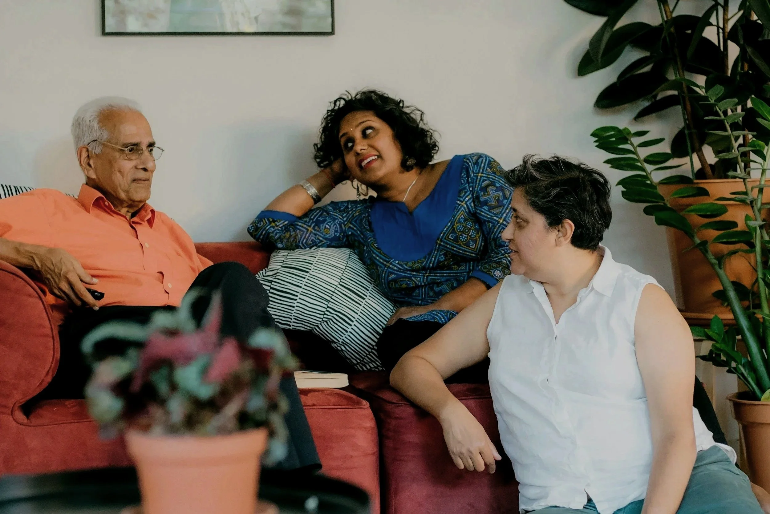 Three people sitting on a red couch having a conversation, with a woman leaning on a pillow and a large potted plant nearby.