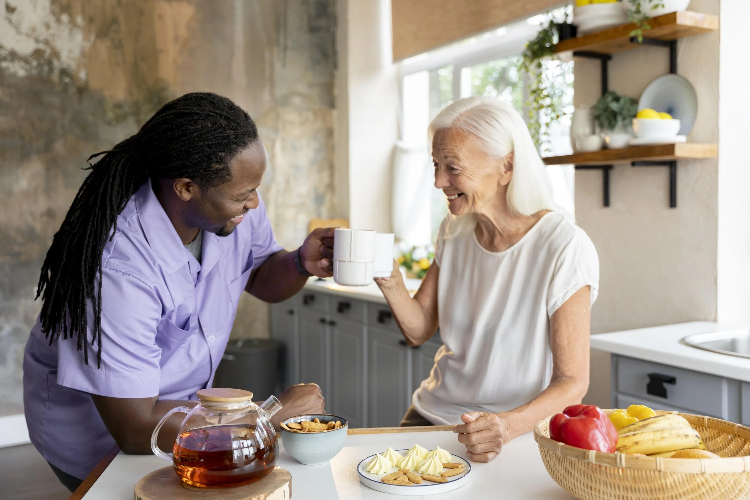 A man with long dreadlocks and a woman with white hair sharing a toast in a kitchen, with snacks and a teapot on the counter.