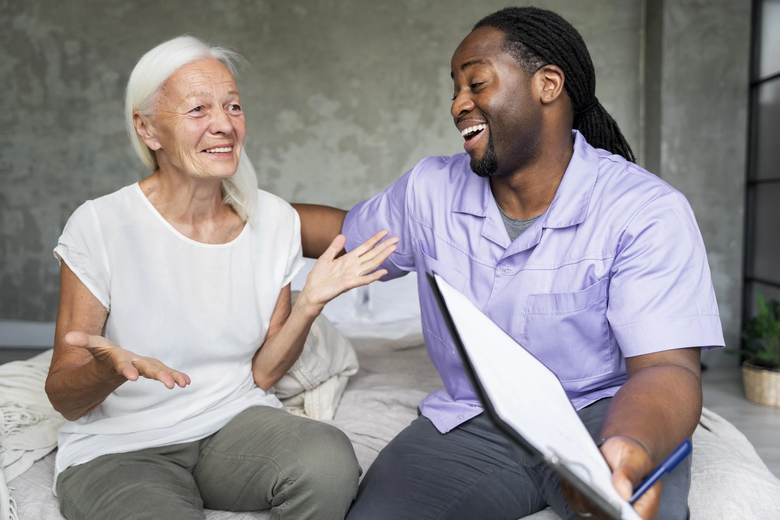 A nurse and an elderly woman are sitting on a bed, smiling, and engaging in a positive conversation in a clinical setting.