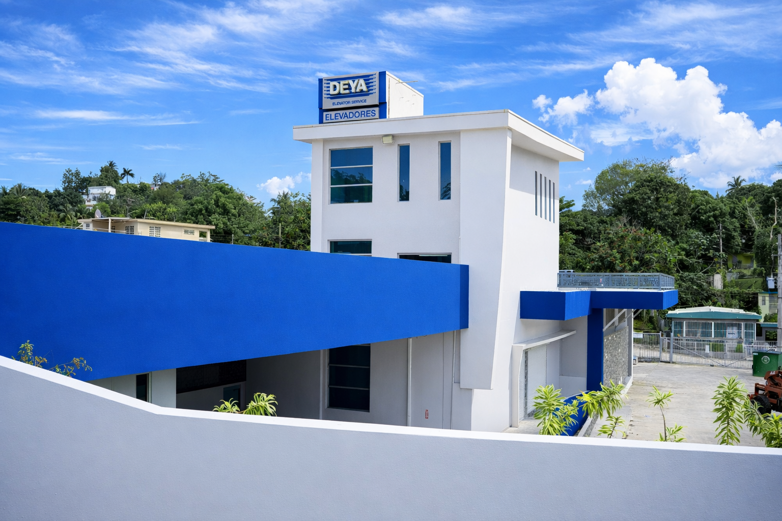 Edificio de color blanco y azul con letrero que dice 'DEYA ELEVATOR SERVICE' en la azotea, y un fondo de cielo azul con nubes y vegetación verde.